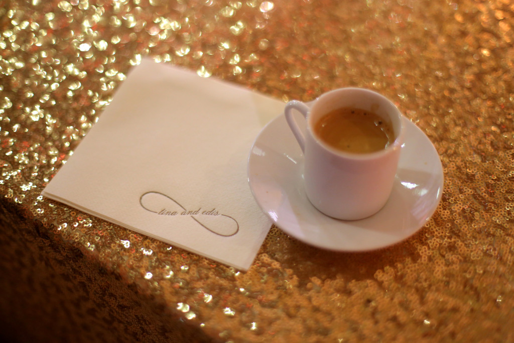single shot of espresso in a white porcelain espresso cup on a saucer sitting on a gold lame tablecloth with a monogrammed napkin