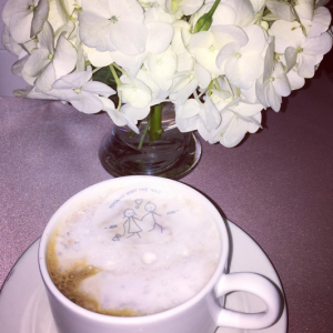 Vase of white flowers and cappuccino in a white coffee cup on a saucer featuring the Original Beverage Topper, an edible disc with a logo on top of the cappuccino foam.
